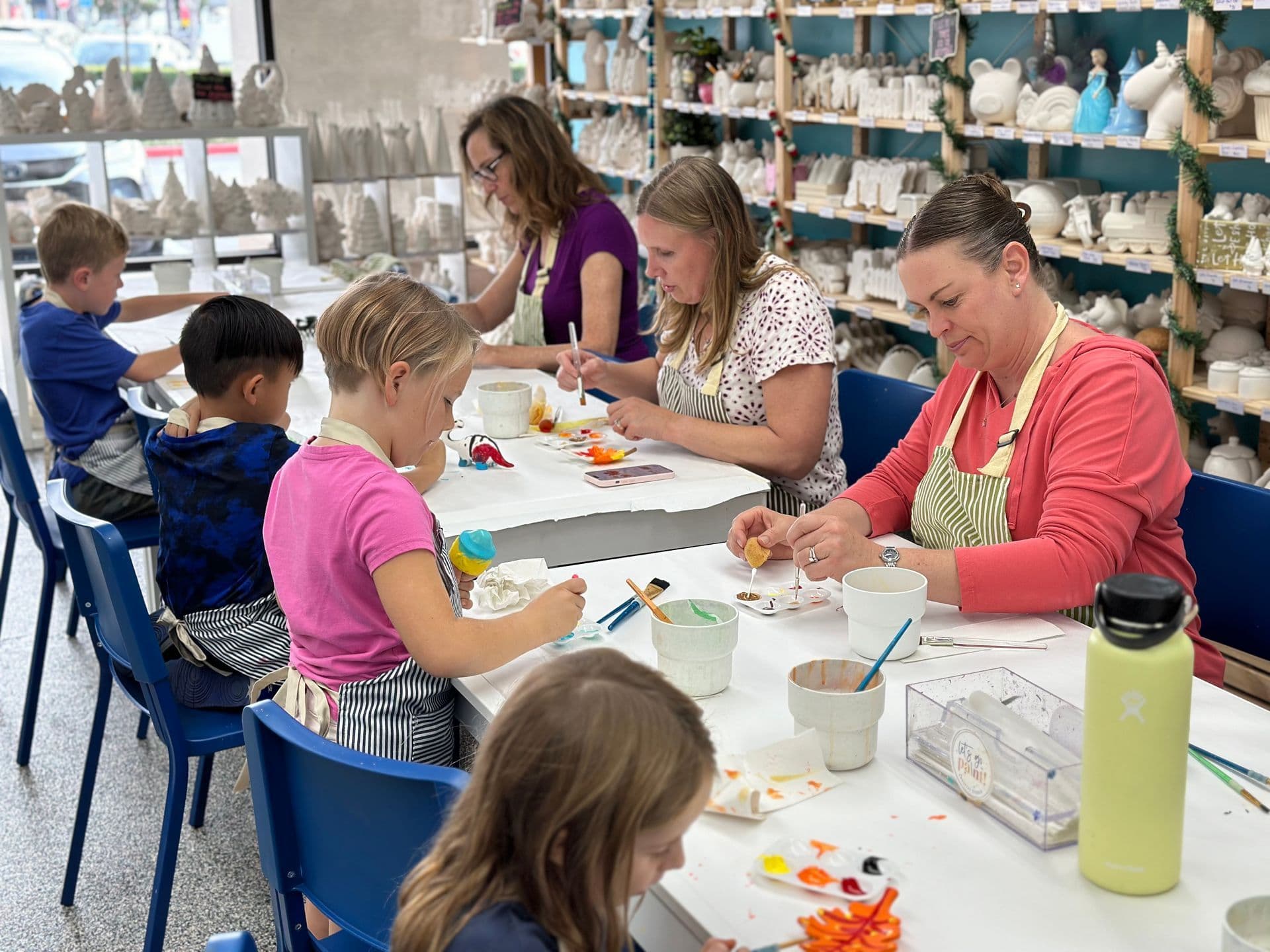 A multigenerational table of kids and a parent painting ceramics together in Faith's Seal Beach studio — pottery shelves in the background, bright colors on the table.